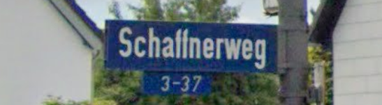 Straßenschild, das mit der Aufschrift Schaffnerweg versehen ist. Im Hintergrund angedeutete sind Häuser und Büsche.